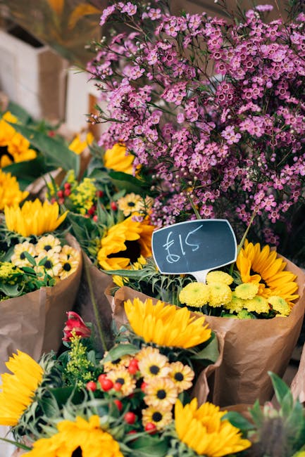 A vibrant floral display featuring a variety of fresh flowers arranged in brown paper wrapping. Prominent bright yellow sunflowers with large, round petals and dark centers are grouped together, accen