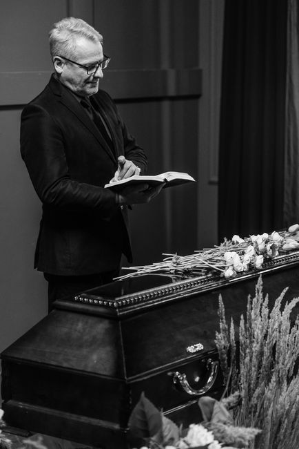 A man with light-colored hair, glasses, and dressed in a dark suit is standing next to a closed casket adorned with a floral arrangement of white roses and greenery. The man is holding a book or condo