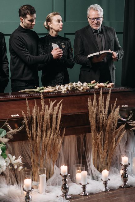 Three individuals stand behind a funeral casket decked with a floral arrangement consisting of white roses, lilies, and greenery, surrounded by tall dried grasses and lit candles in silver holders. Th