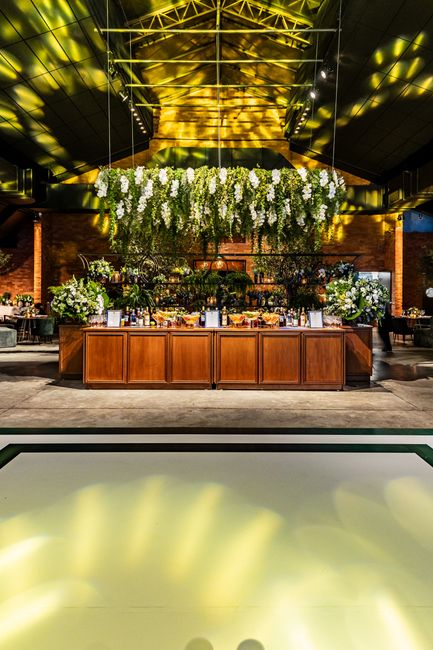 A large floral arrangement comprised of cascading white flowers, likely including white wisteria and roses, is suspended above a wooden bar or buffet table within an industrial-style venue. The arrang
