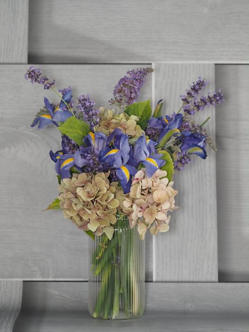 A floral arrangement featuring blue and yellow iris flowers, soft pink and cream hydrangeas, and sprigs of purple lavender, all set in a clear glass vase. The bouquet is arranged with vibrant blooms a